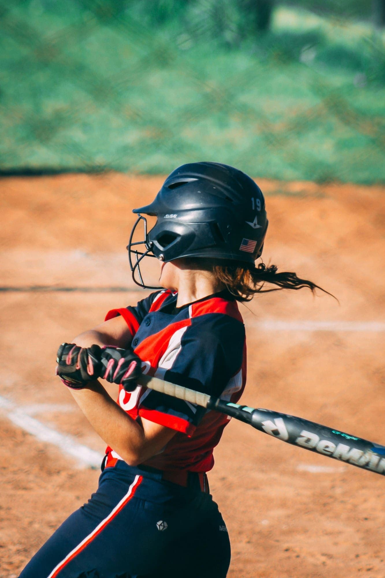 Softball hitter finishing a swing, ponytail flying under the helmet